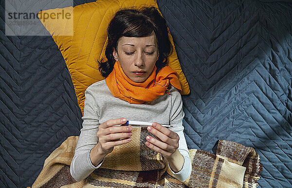 Sick woman checking temperature on thermometer lying on bed at home