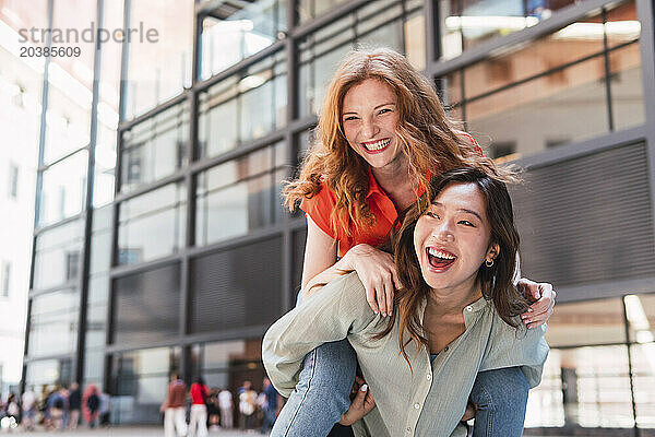 Happy woman piggybacking friend in front of building