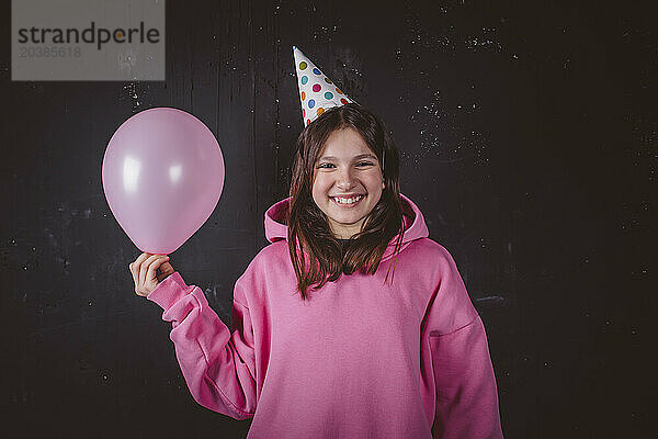 Cheerful girl in hat holding pink balloon at birthday party in front of black background