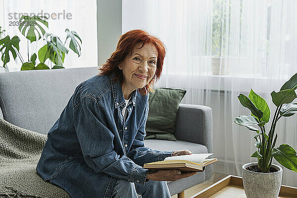 Smiling retired senior woman reading book on sofa at home