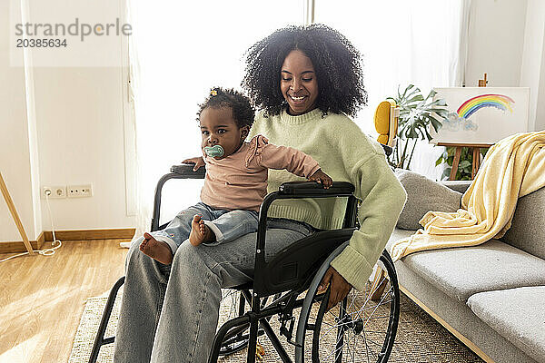 Cute baby girl sitting on mother with disability in wheelchair at home