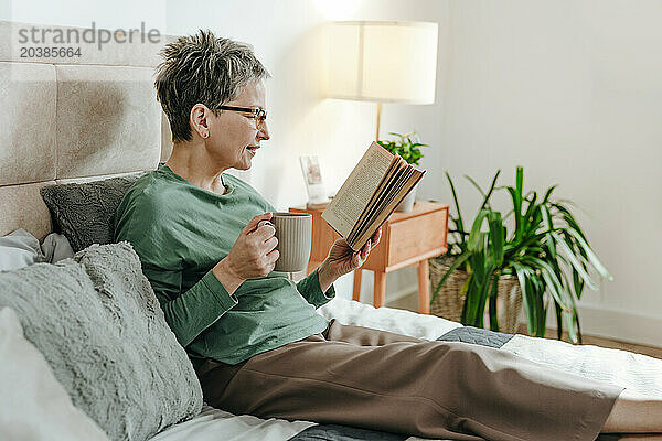 Woman holding mug and reading book on bed at home