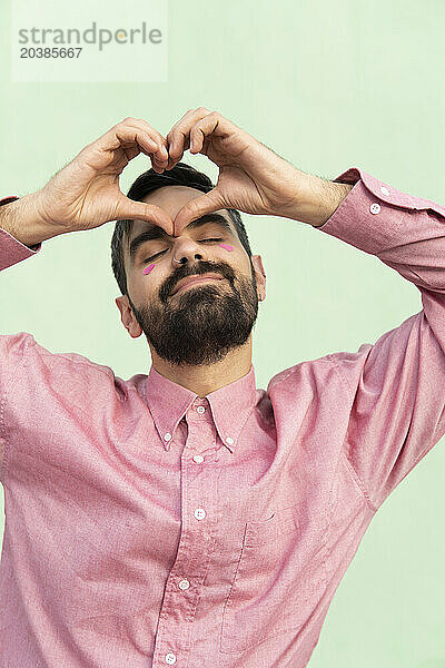 Smiling man gesturing heart shape against mint green background