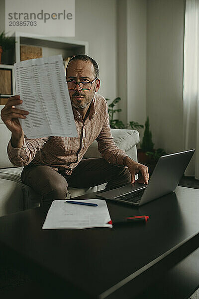 Mature freelancer reading document near laptop at home office