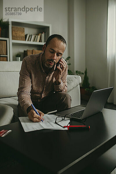 Freelancer talking through smart phone and writing on paper at home office