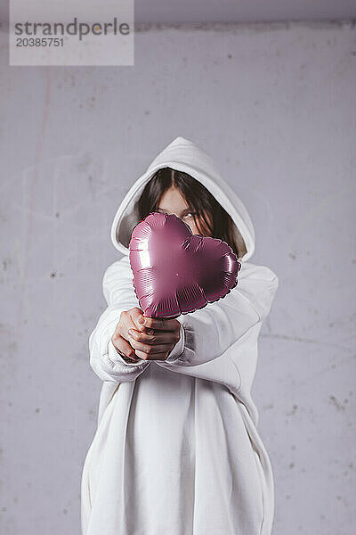 Girl covering face with heart shaped pink balloon in front of gray cement background