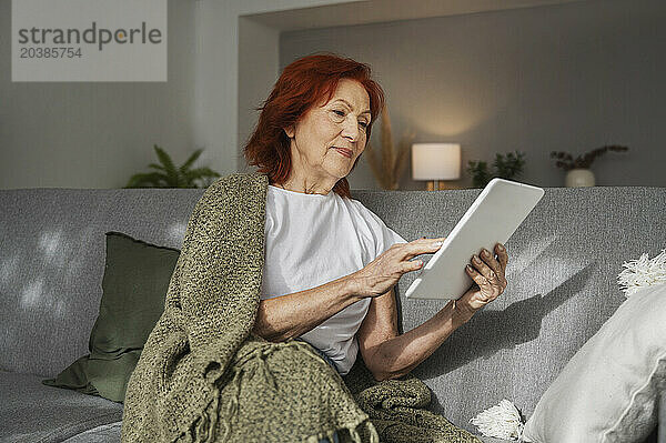 Redhead retired woman with digital tablet sitting on sofa at home