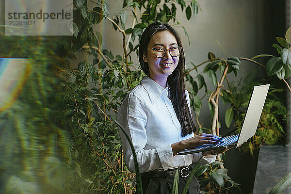 Happy young businesswoman standing with laptop in office