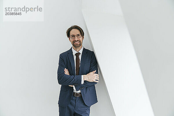 Smiling businessman leaning on white wall with arms crossed at office