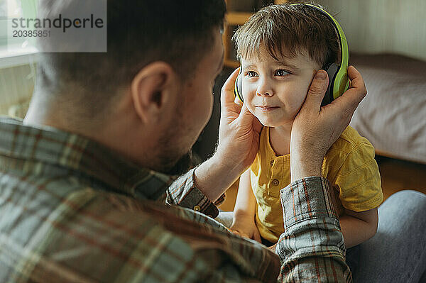 Father with son wearing wireless headphones and sitting on lap at home