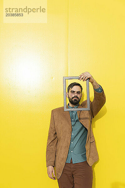 Man looking through picture frame in front of yellow wall