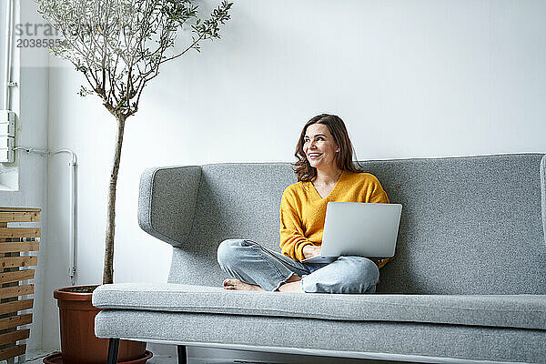 Happy woman sitting cross-legged on sofa with laptop in living room at home