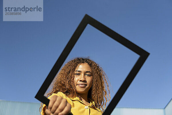 Confident young woman looking through picture frame under clear blue sky