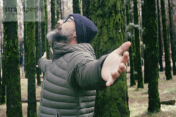 Carefree man with arms outstretched leaning on tree trunk in forest