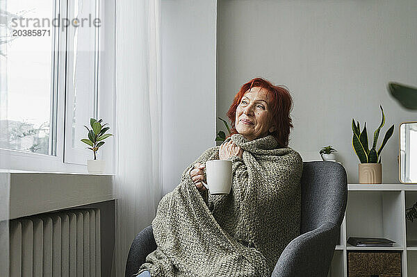 Redhead senior woman sitting in armchair with mug of tea by radiator at home