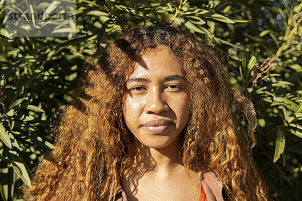 Young woman with curly hair in front of plants on sunny day