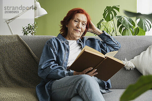 Relaxed senior woman reading book sitting on sofa at home