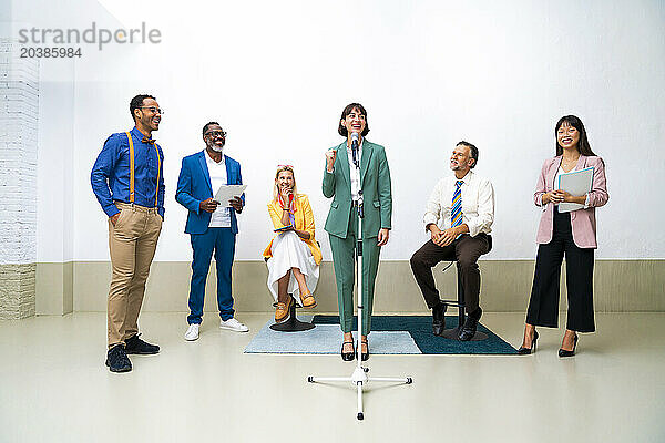 Happy public speaker amidst colleagues near wall at office