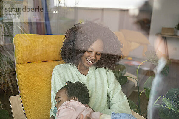 Smiling mother sitting on chair with baby girl seen through window glass