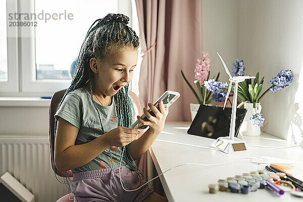 Surprised girl holding smart phone near wind turbine model at table