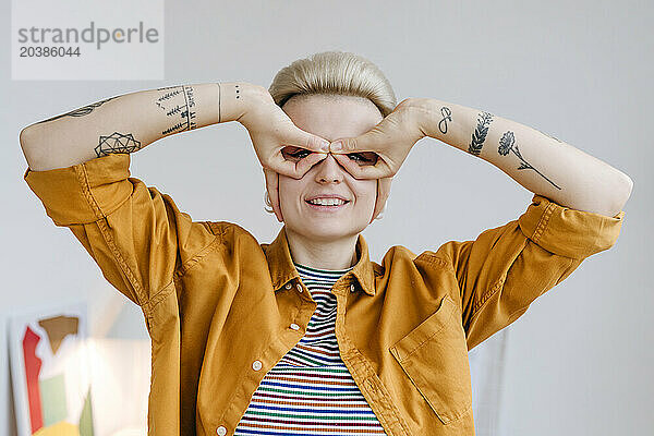 Smiling woman holding fingers to eyes like glasses near white wall