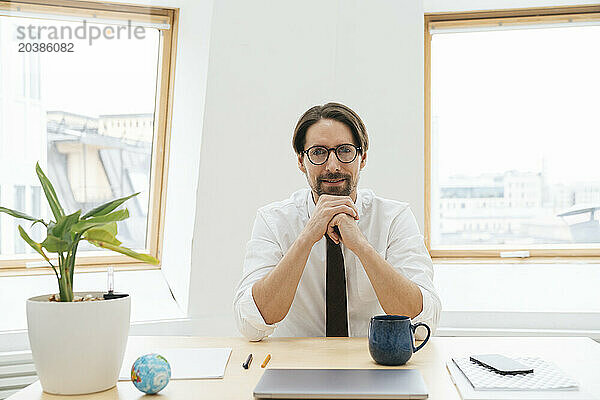 Smiling businessman sitting with laptop and coffee cup at desk in window