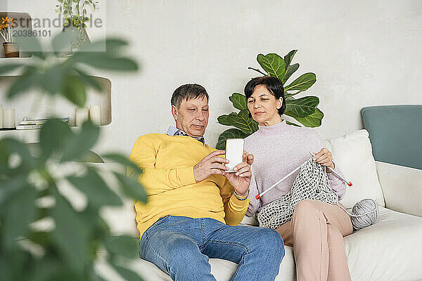 Man showing smart phone to woman knitting on sofa at home