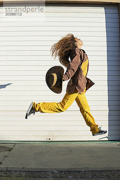 Excited young woman with cowboy hat jumping on footpath by white shutter