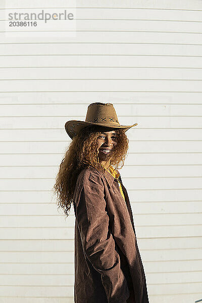 Smiling young woman with curly hair wearing jacket and cowboy hat on sunny day