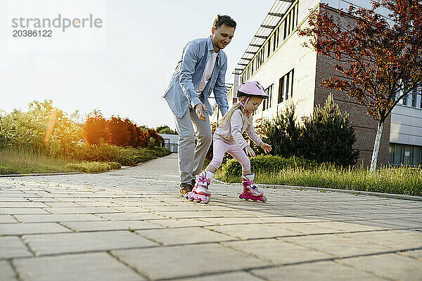 Father assisting daughter roller skating on footpath