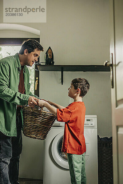 Son helping father in holding basket near washing machine at home