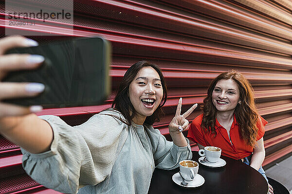 Woman making peace sign and taking selfie with friend through smart phone