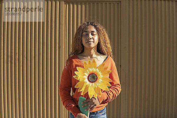 Young woman with eyes closed holding artificial sunflower in front of metal wall