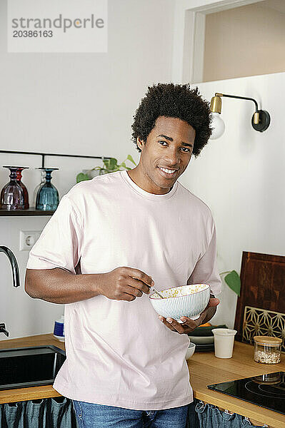 Smiling man standing in kitchen eating breakfast from bowl at home