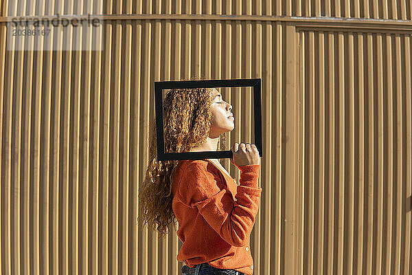 Young woman with eyes closed holding picture frame standing in front of metal wall