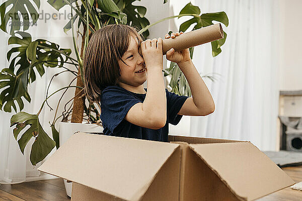 Happy boy sitting in box and looking through cardboard telescope at home
