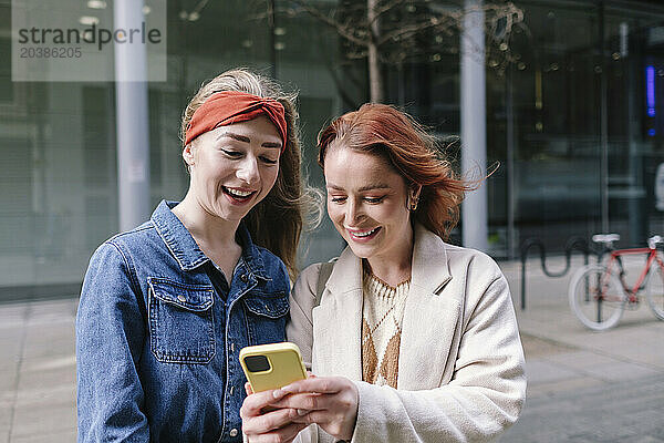 Happy woman with friend using smart phone in front of building