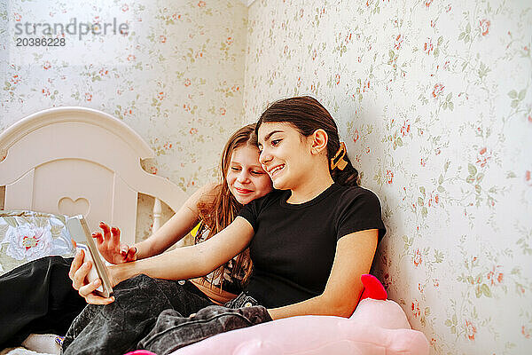 Smiling girl taking selfie with friend sitting near wall at home