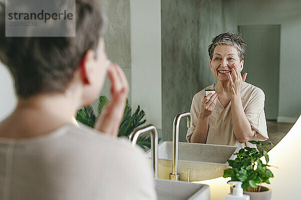 Smiling woman applying moisturizer on face looking in mirror at home