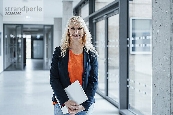 Happy mature businesswoman holding laptop and standing in office corridor
