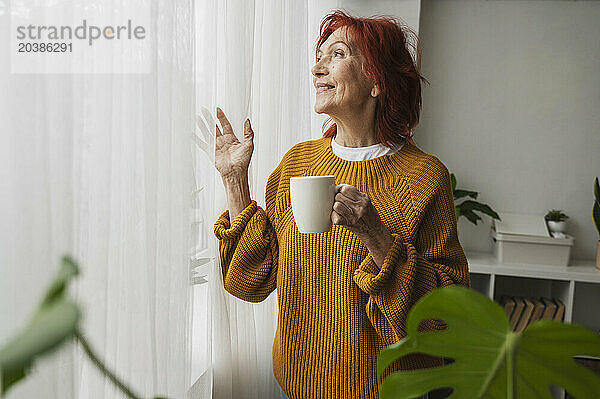 Retired senior woman standing with tea cup by window day dreaming at home