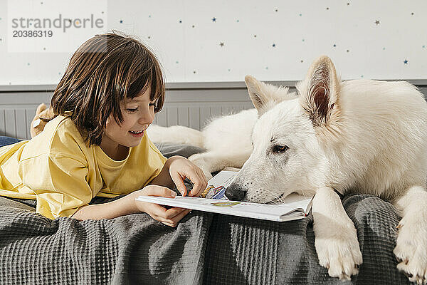 Smiling boy reading book with white Swiss shepherd dog on bed