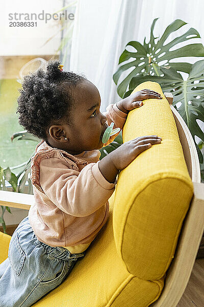 Cute baby girl with pacifier leaning on chair at home