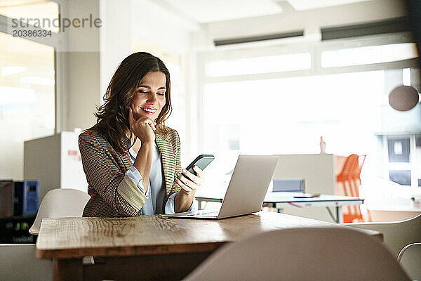 Smiling businesswoman using mobile phone at laptop desk in office