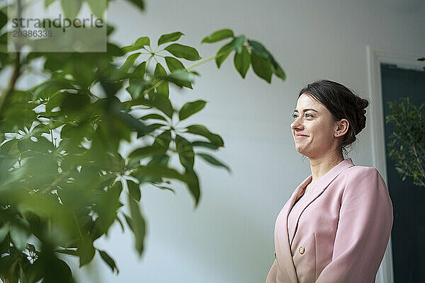 Smiling businesswoman wearing blazer standing near plant at home