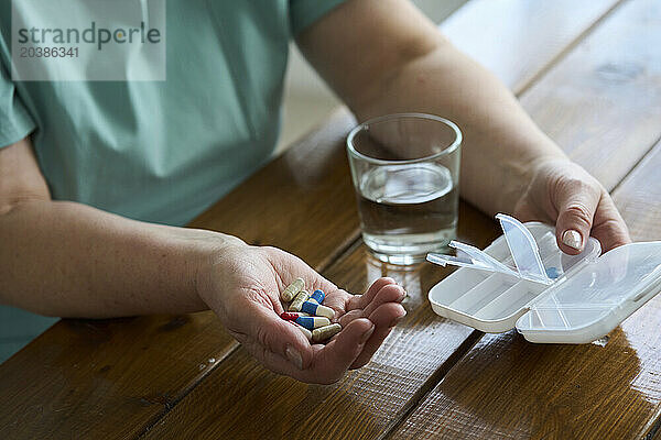 Woman holding capsules in hand by glass of water on table at home