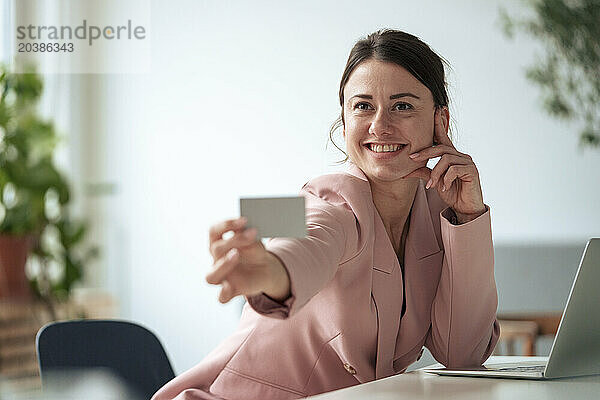 Smiling businesswoman sitting with laptop at table showing business card working at home