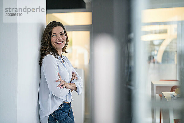 Smiling confident businesswoman with arms crossed leaning on wall in office