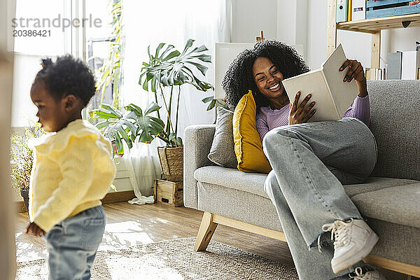 Happy woman reading book reclining on sofa near daughter standing in living room at home