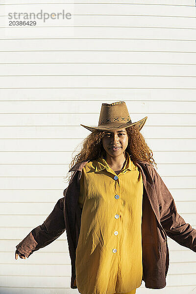 Confident young woman with curly hair wearing jacket and cowboy hat on sunny day